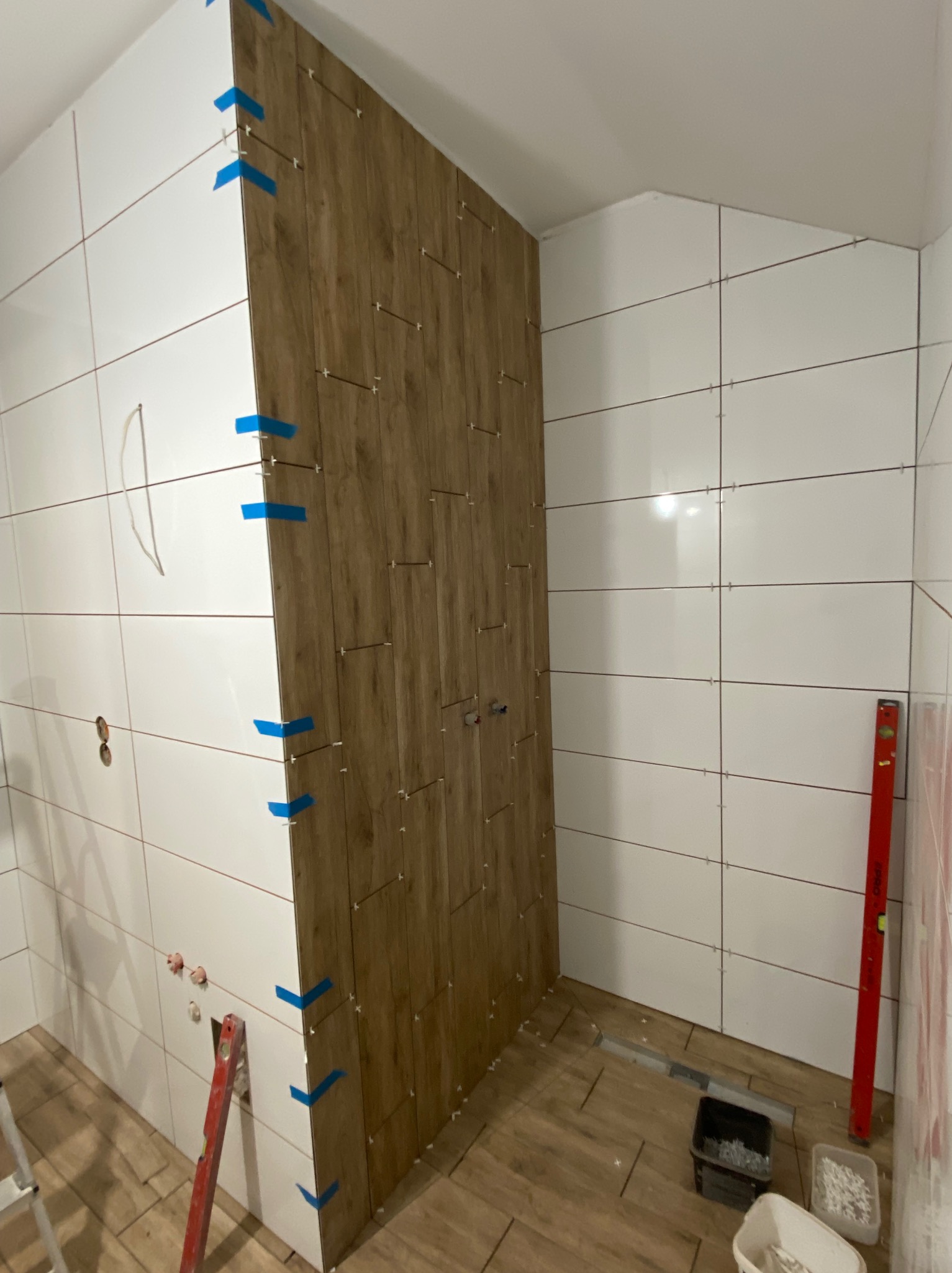 Montaż płytek w łazience: białe, prostokątne płytki na ścianie kontrastują z pionowym pasem płytek imitujących drewno, zabezpieczonych niebieską taśmą; widoczne krzyżyki dystansowe, poziomica...