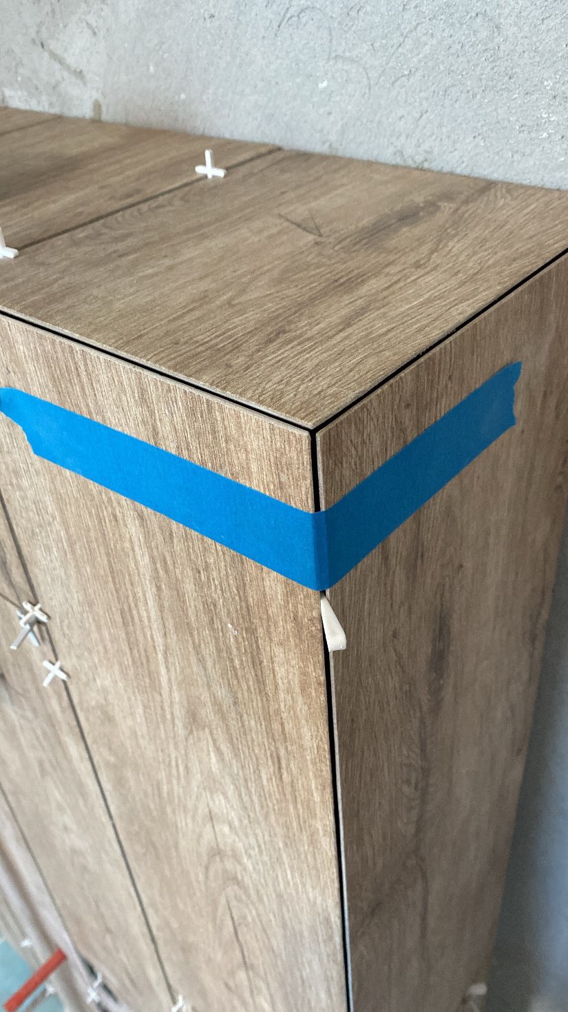 Fragment ściany z drewnopodobnymi płytkami w trakcie montażu, zabezpieczony niebieską taśmą malarską i krzyżykami dystansowymi, surowy beton w tle.