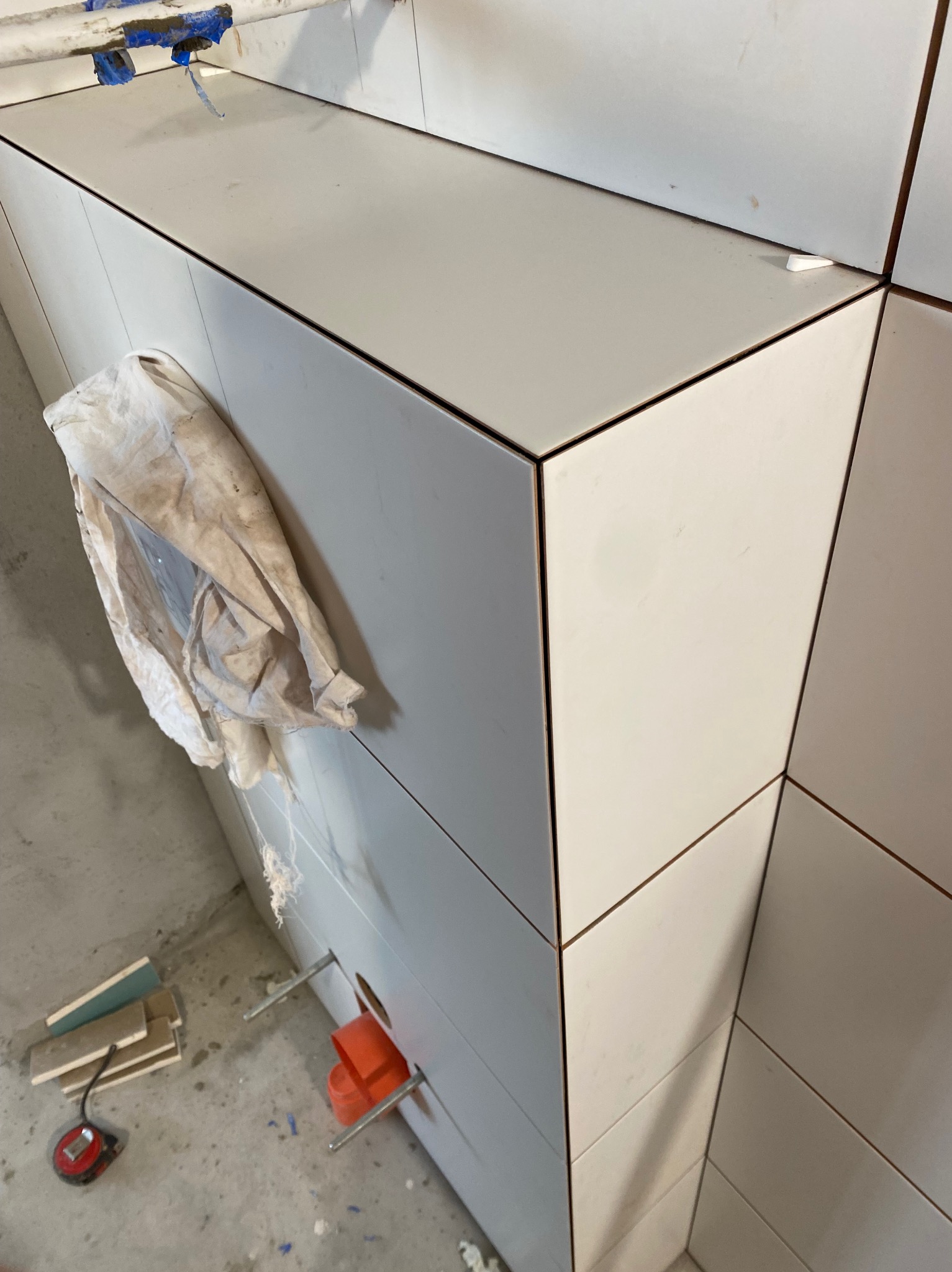 Białe płytki ułożone na ścianie w łazience, widoczne narzędzia i brudna szmatka, fragment rury i przymiaru na podłodze, etap prac wykończeniowych.