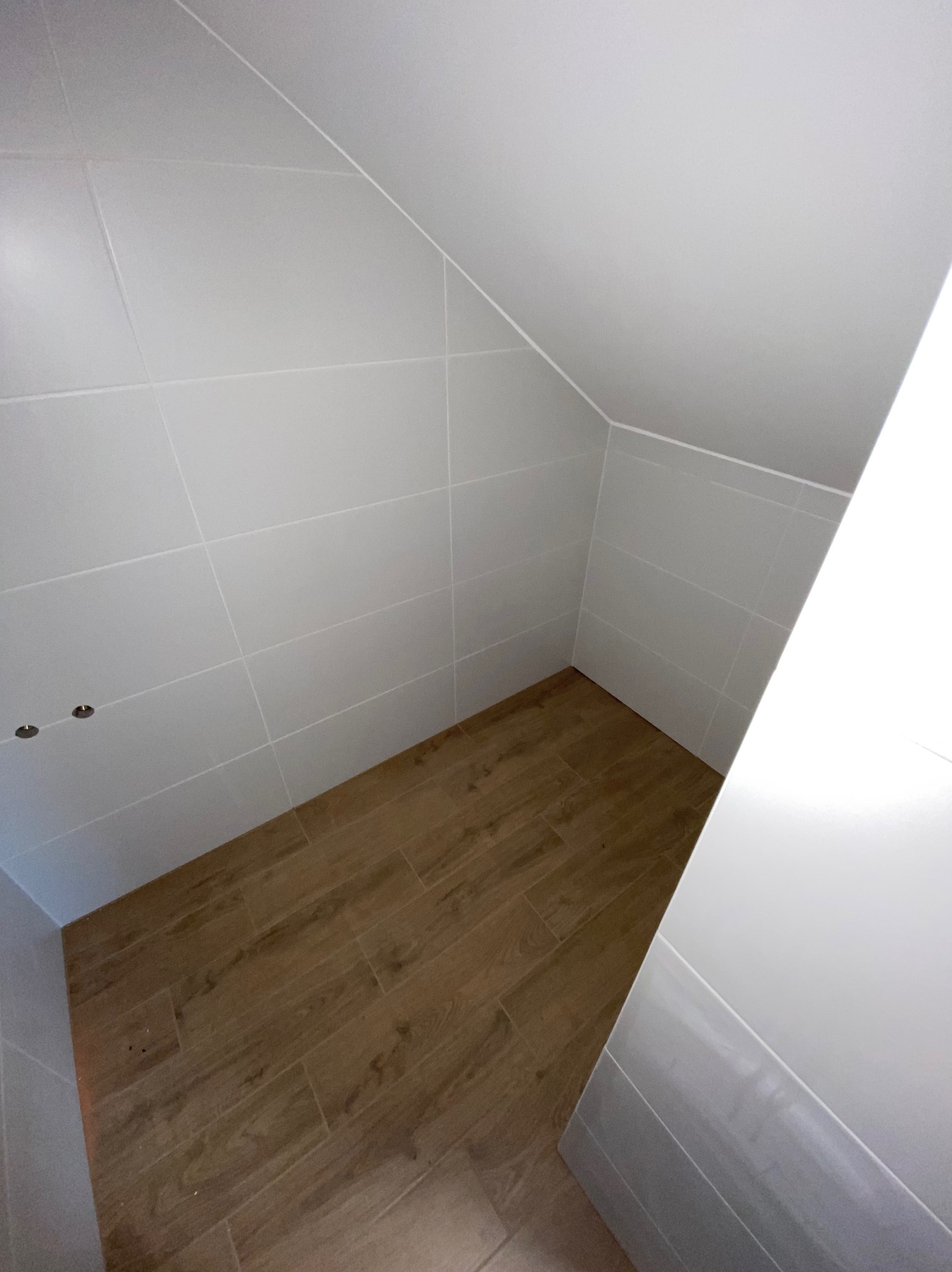 Wykończona łazienka na poddaszu ze skośnym sufitem, wyłożona białymi płytkami, podłoga z paneli imitujących drewno, widoczne dwa otwory na oświetlenie w suficie.