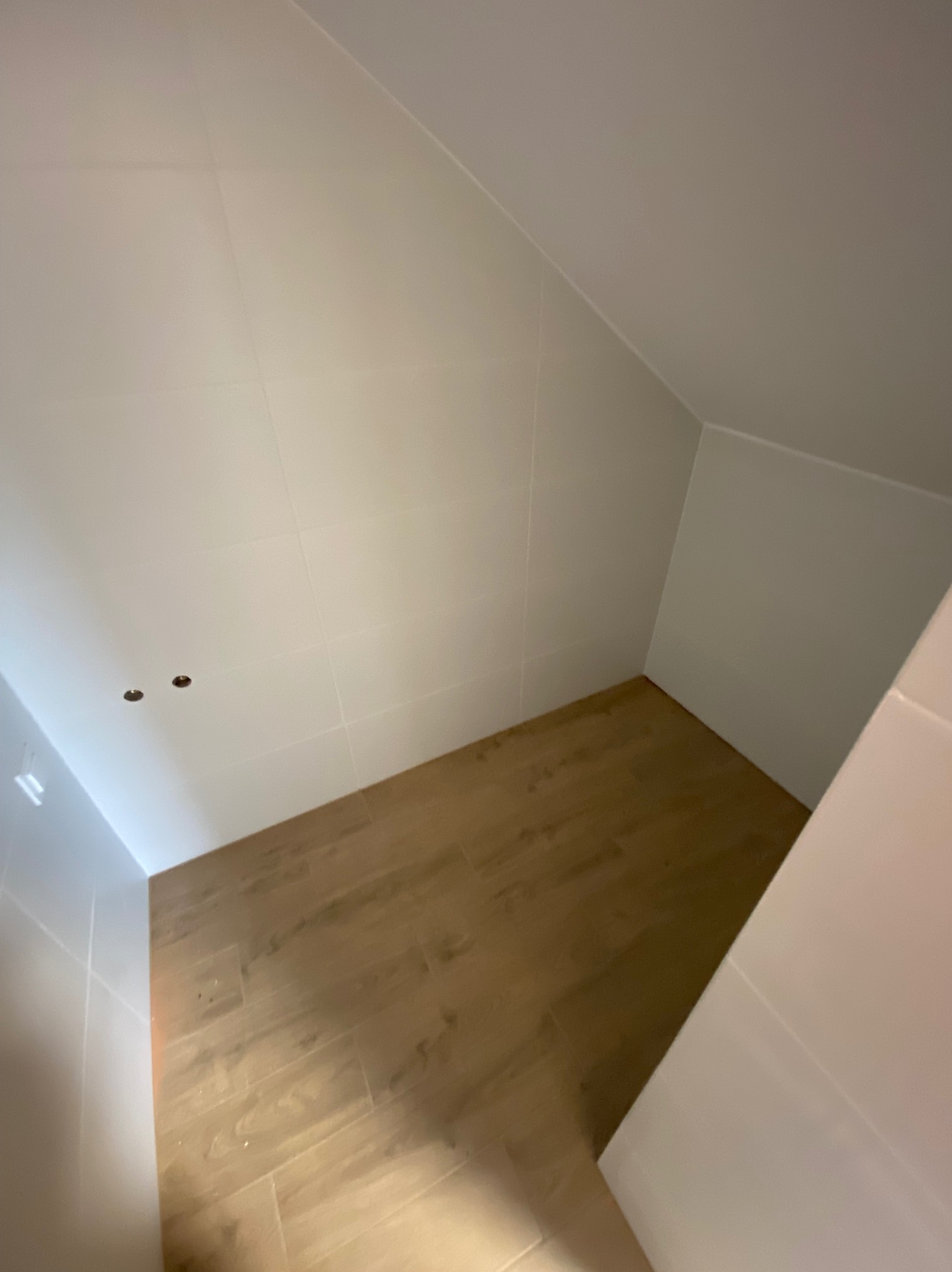 Mała łazienka na poddaszu z jasnymi płytkami na ścianach i podłogą imitującą drewno, widoczne otwory na oświetlenie w suficie.