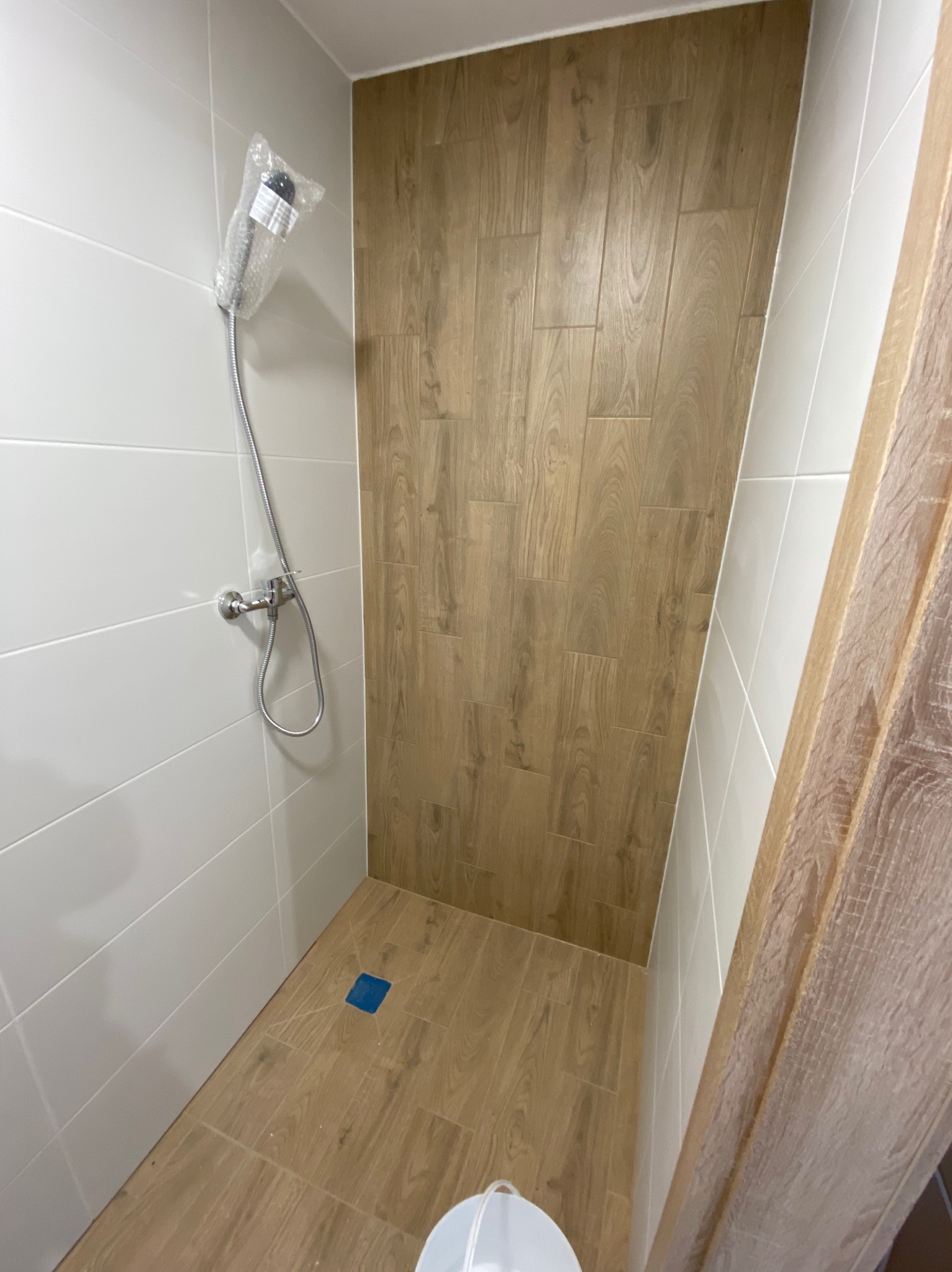 Nowa kabina prysznicowa z drewnopodobnymi płytkami ściennymi, białe płytki na pozostałych ścianach, niebieski odpływ podłogowy i zapakowana słuchawka prysznicowa.