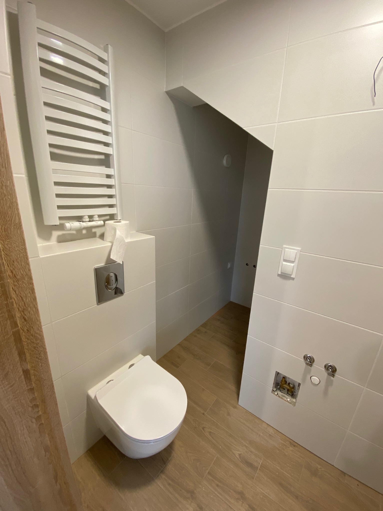 Nowoczesna, biała łazienka z podwieszaną toaletą, grzejnikiem drabinkowym, drewnopodobną podłogą i przygotowanym miejscem na baterię umywalkową.
