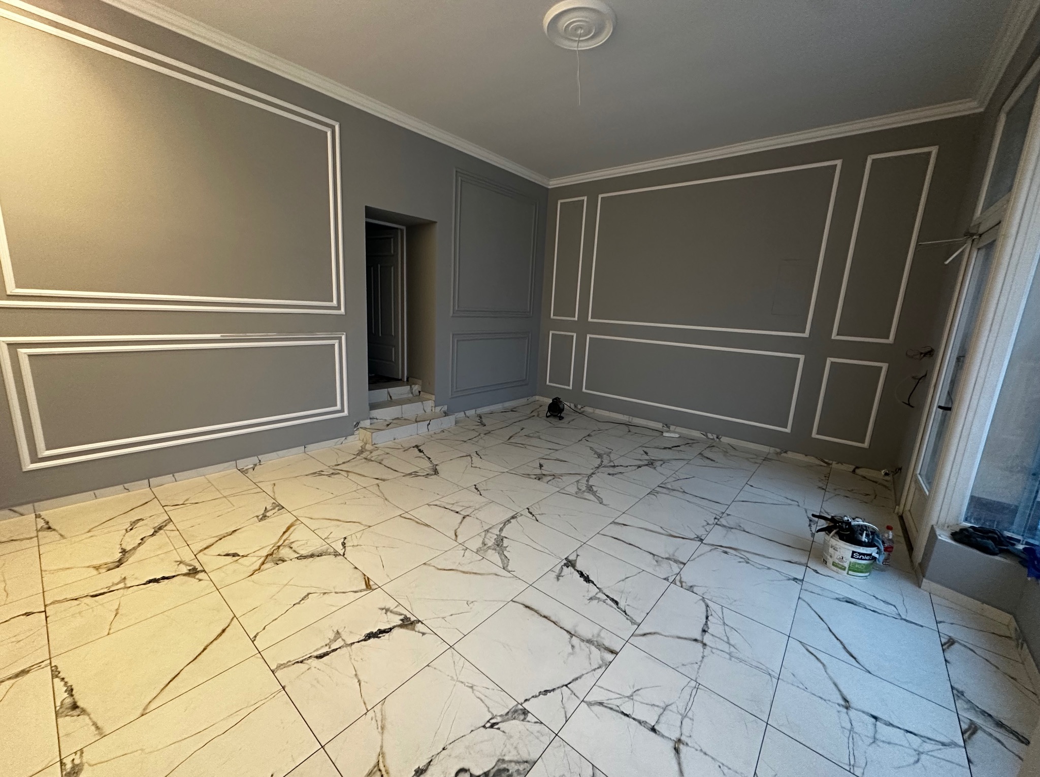 Szare ściany z białymi sztukateriami w pustym pokoju z marmurową podłogą. Widoczny fragment drzwi i wiadro z farbą. Nowoczesne wykończenie wnętrza.