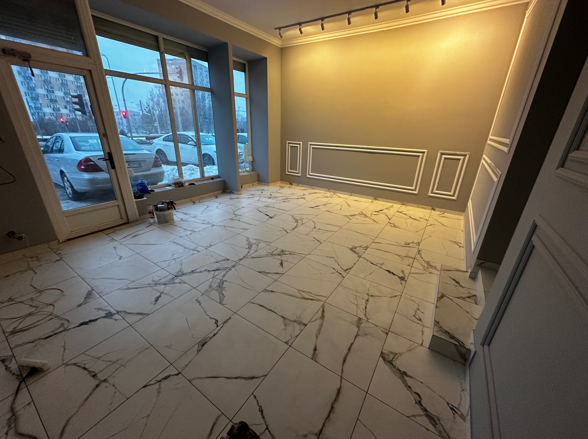 Wnętrze po remoncie: marmurowe płytki podłogowe, szare ściany z białymi dekoracyjnymi listwami, duże okna z widokiem na ulicę. Widoczne narzędzia pracy.