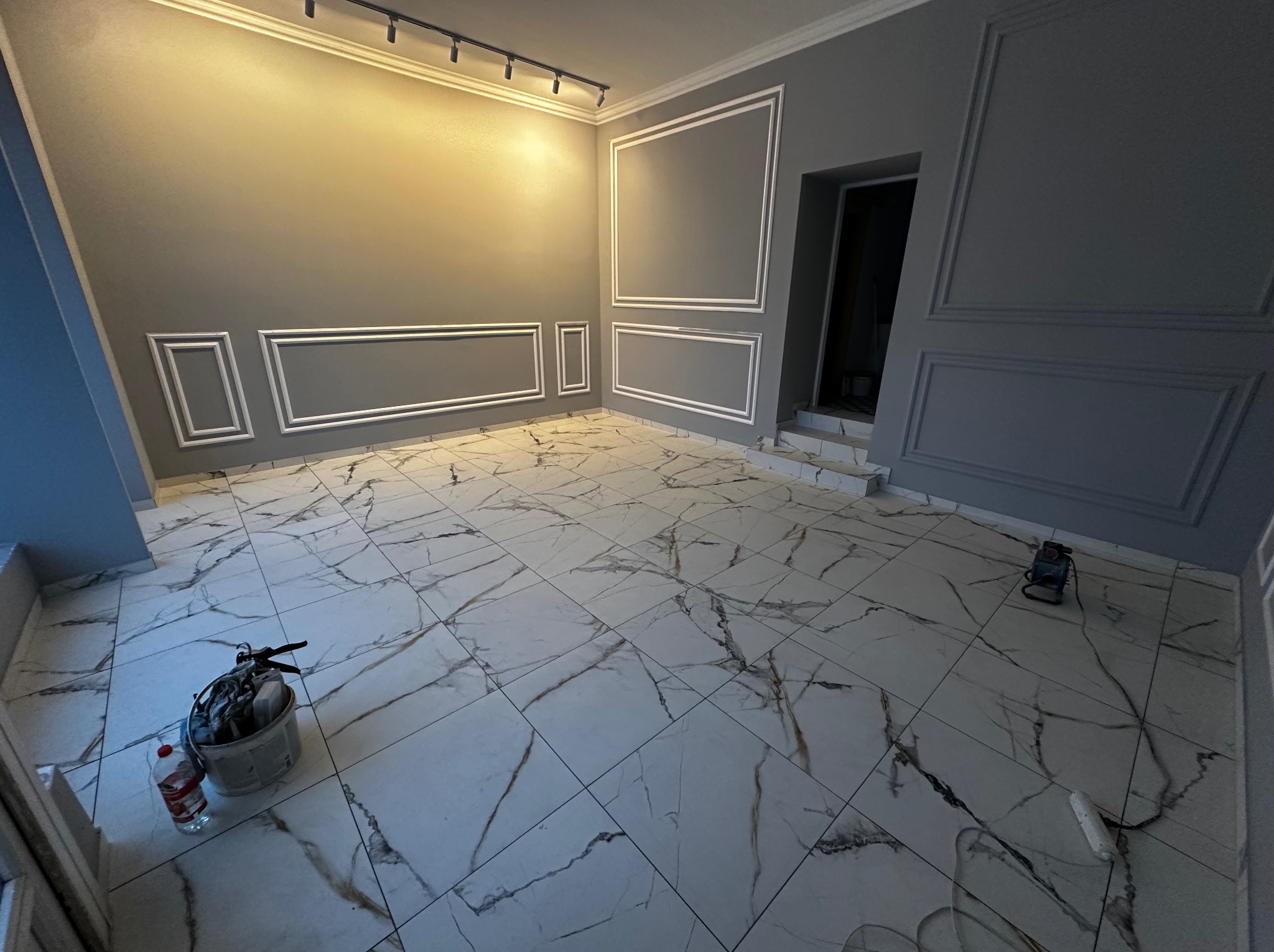 Szare wnętrze po remoncie z białymi sztukateriami na ścianach i marmurową podłogą. Widoczne narzędzia malarskie i oświetlenie sufitowe.