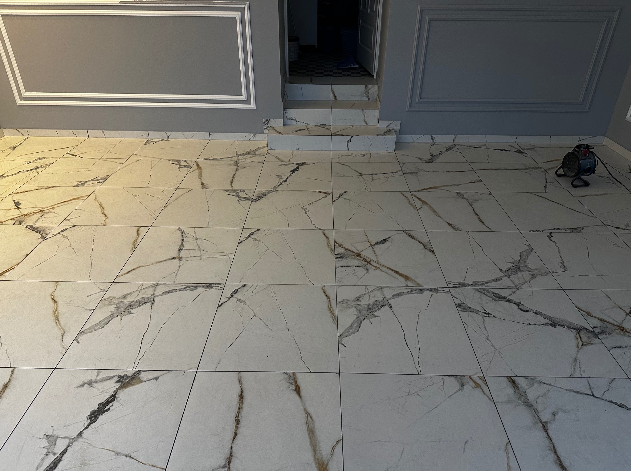 Ułożone białe płytki podłogowe z szarymi i złotymi żyłami, dopasowane do schodów i listew przypodłogowych. Szare ściany z białymi dekoracyjnymi ramami.