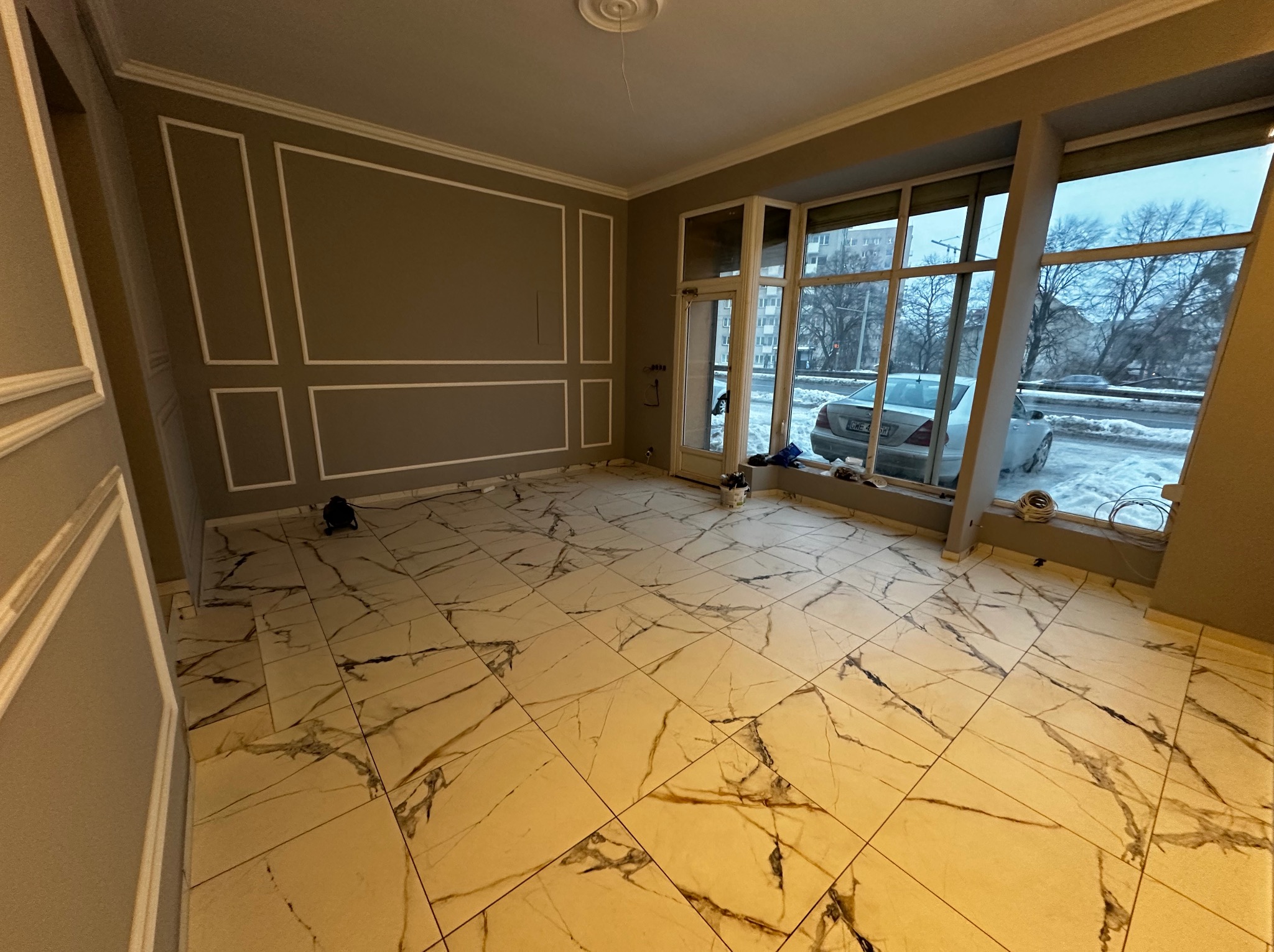Pokój po remoncie z marmurową podłogą i dekoracyjnymi listwami ściennymi. Duże okno z widokiem na zimowy krajobraz. Widoczne narzędzia i akcesoria remontowe.