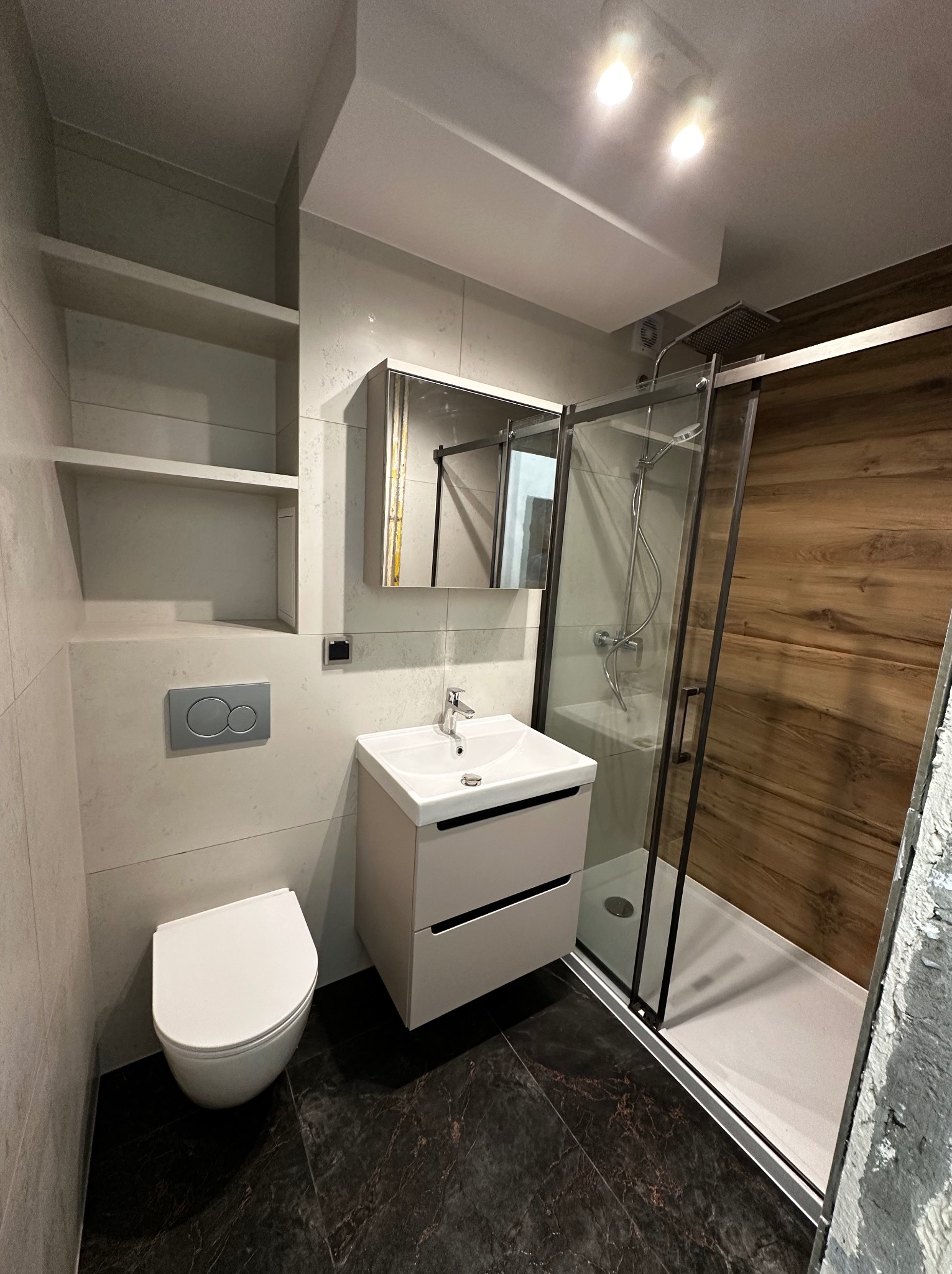 Nowoczesna łazienka z kabiną prysznicową typu walk-in, umywalką z szafką, podwieszaną toaletą i półkami wnękowymi. Wykończenie w odcieniach szarości i drewna.