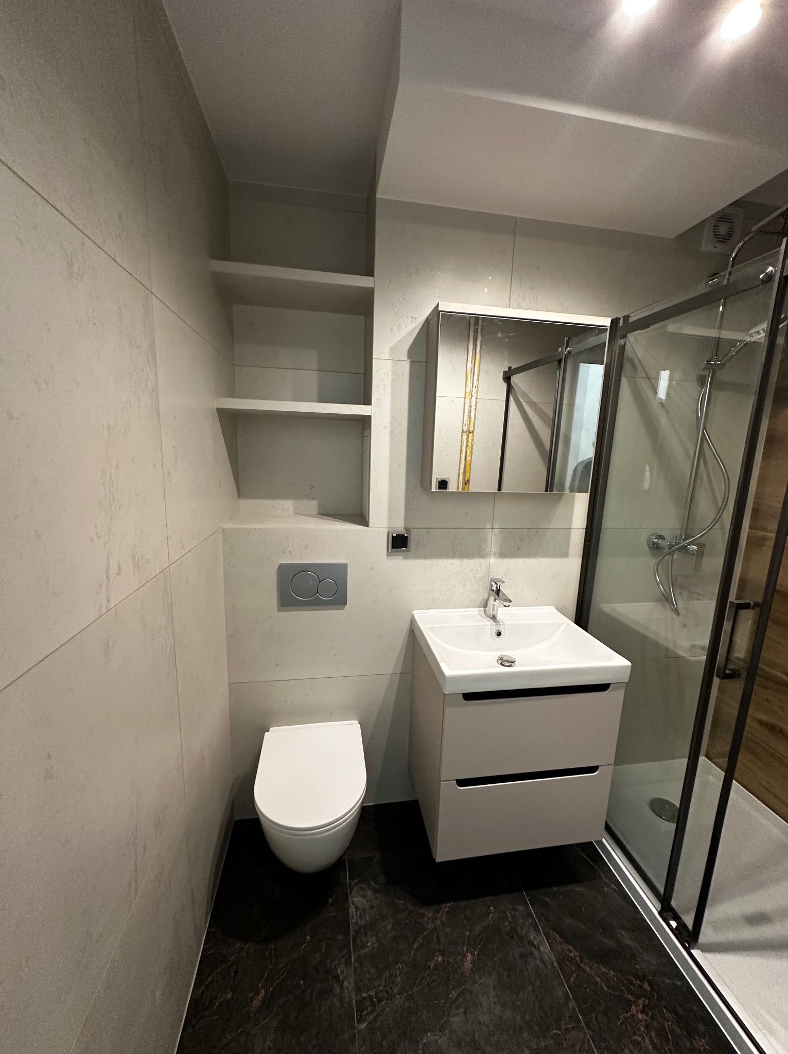 Nowoczesna łazienka po remoncie: kabina prysznicowa z czarną ramą, biała umywalka z szafką, podwieszana toaleta i ciemna marmurowa podłoga. Minimalistyczny design.