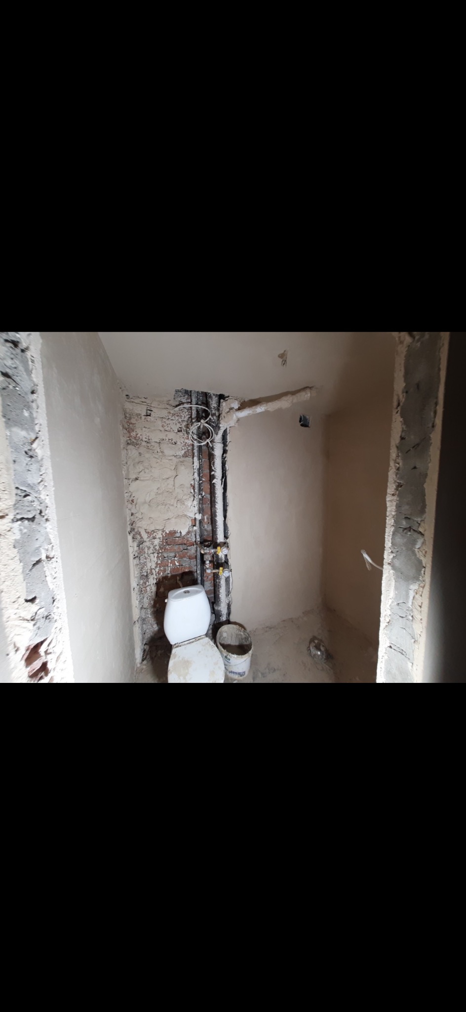 Łazienka w trakcie remontu: widoczne rury, częściowo otynkowane ściany, toaleta i wiadro. Surowy stan przed wykończeniem.
