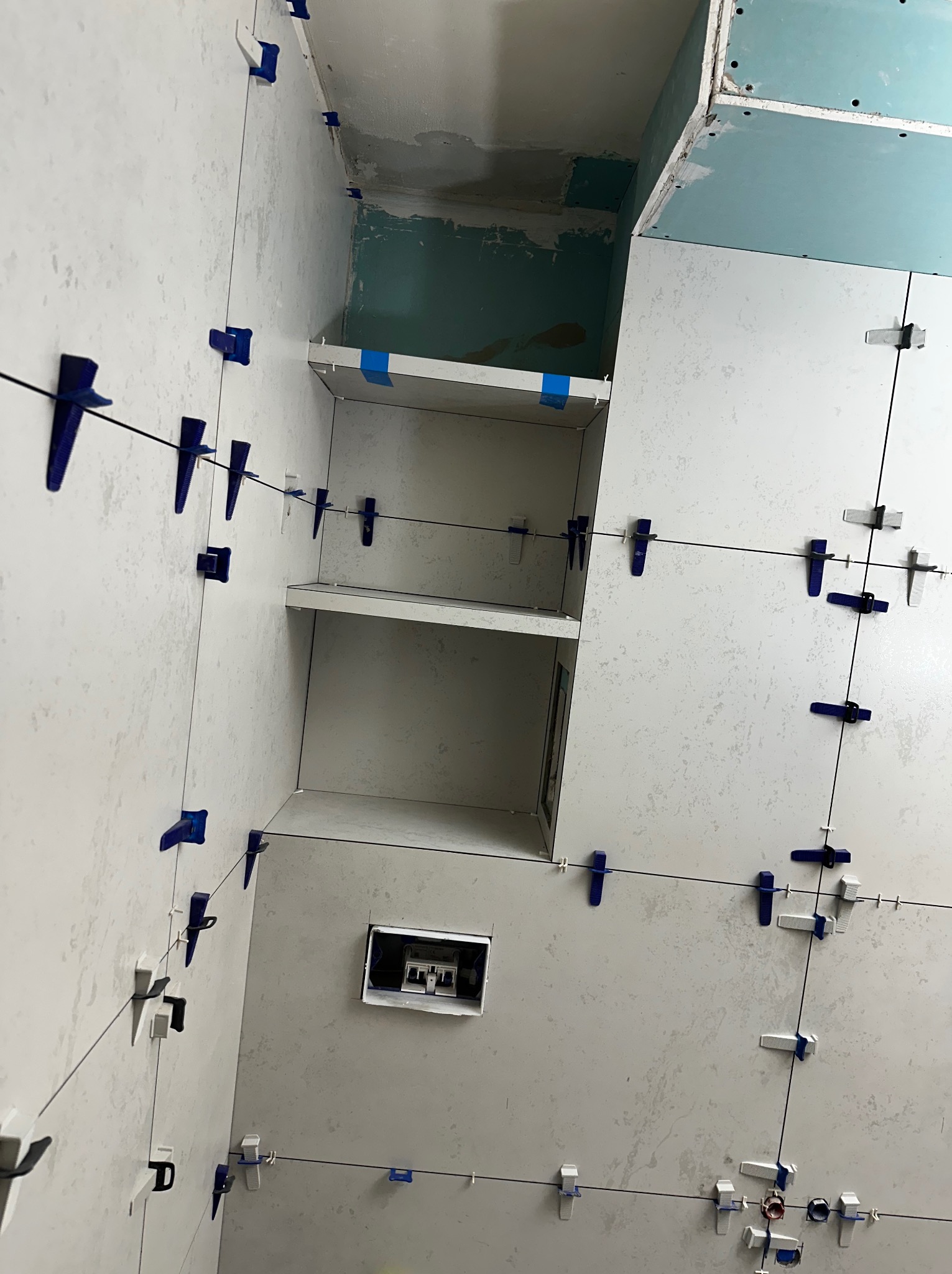 Białe płytki ścienne z niebieskimi klipsami poziomującymi w trakcie montażu w łazience. Widoczne półki wnękowe i sufit z płyt kartonowo-gipsowych.