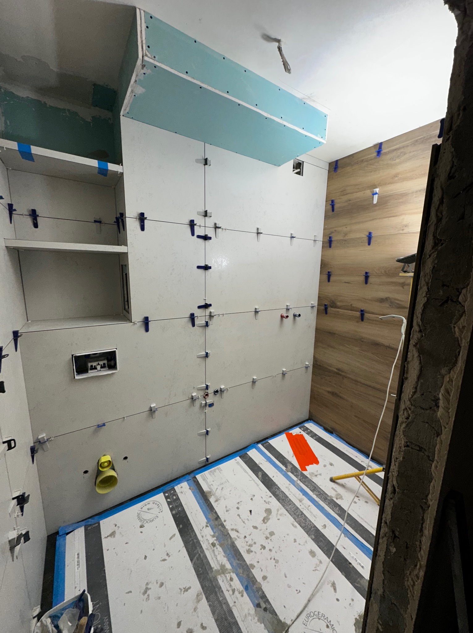 Wnętrze łazienki w trakcie prac wykończeniowych: układanie płytek na ścianach z użyciem klipsów poziomujących, widoczna zabudowa z płyty gipsowej i fragment podłogi z ułożonymi płytkami.