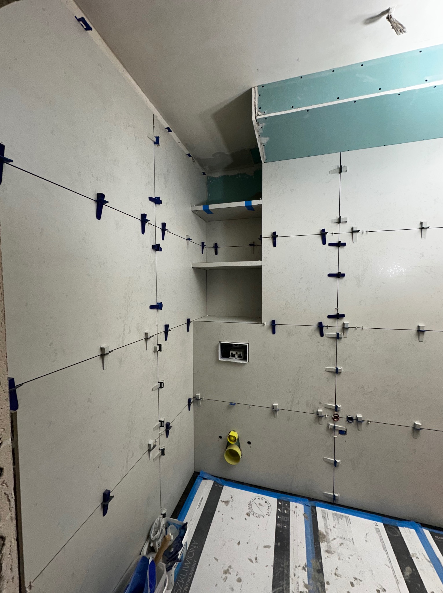 Wnętrze łazienki w trakcie wykańczania: białe płytki z niebieskimi krzyżykami dystansowymi, widoczne półki i fragment płyty karton-gips na suficie. Prace glazurnicze w toku.