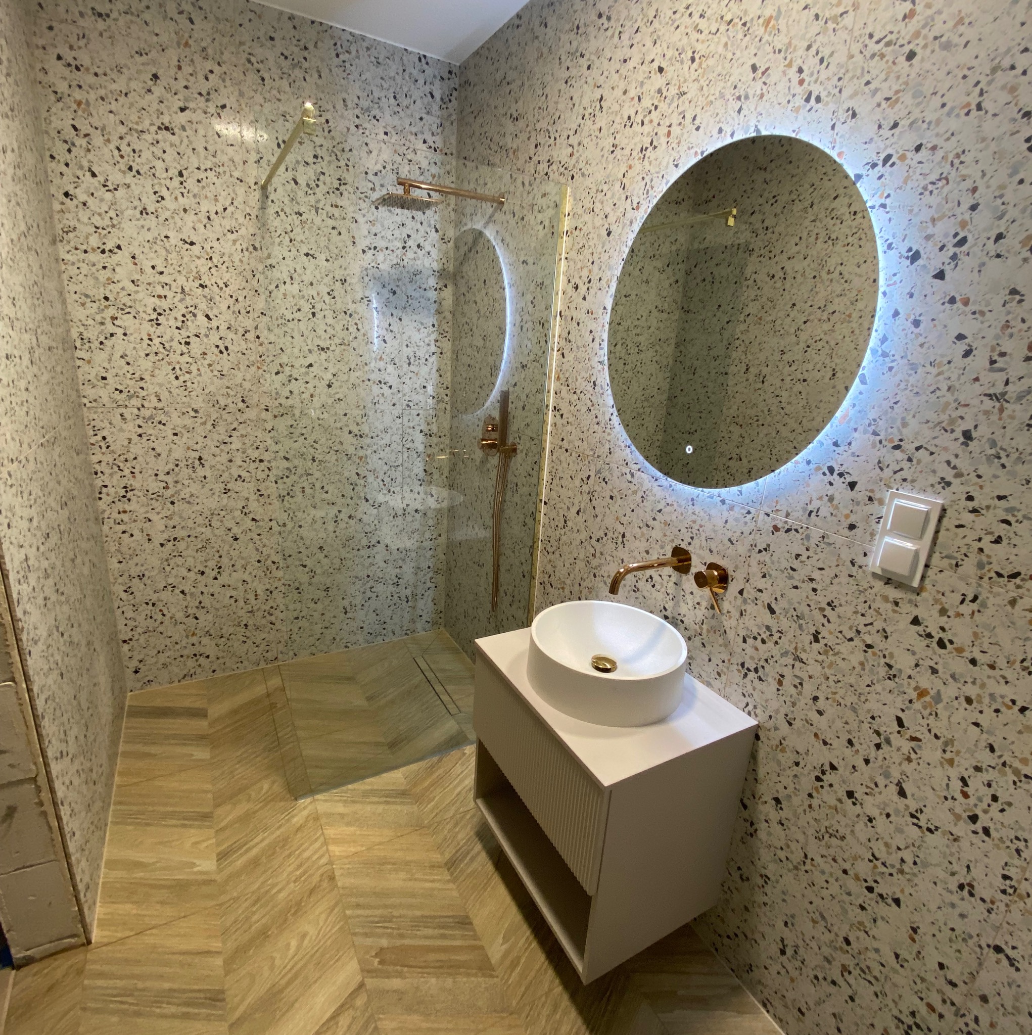 Nowoczesna łazienka z mozaikową ścianą, okrągłym lustrem z podświetleniem LED, białą umywalką nablatową i złotą armaturą. Parkiet ułożony w jodełkę.