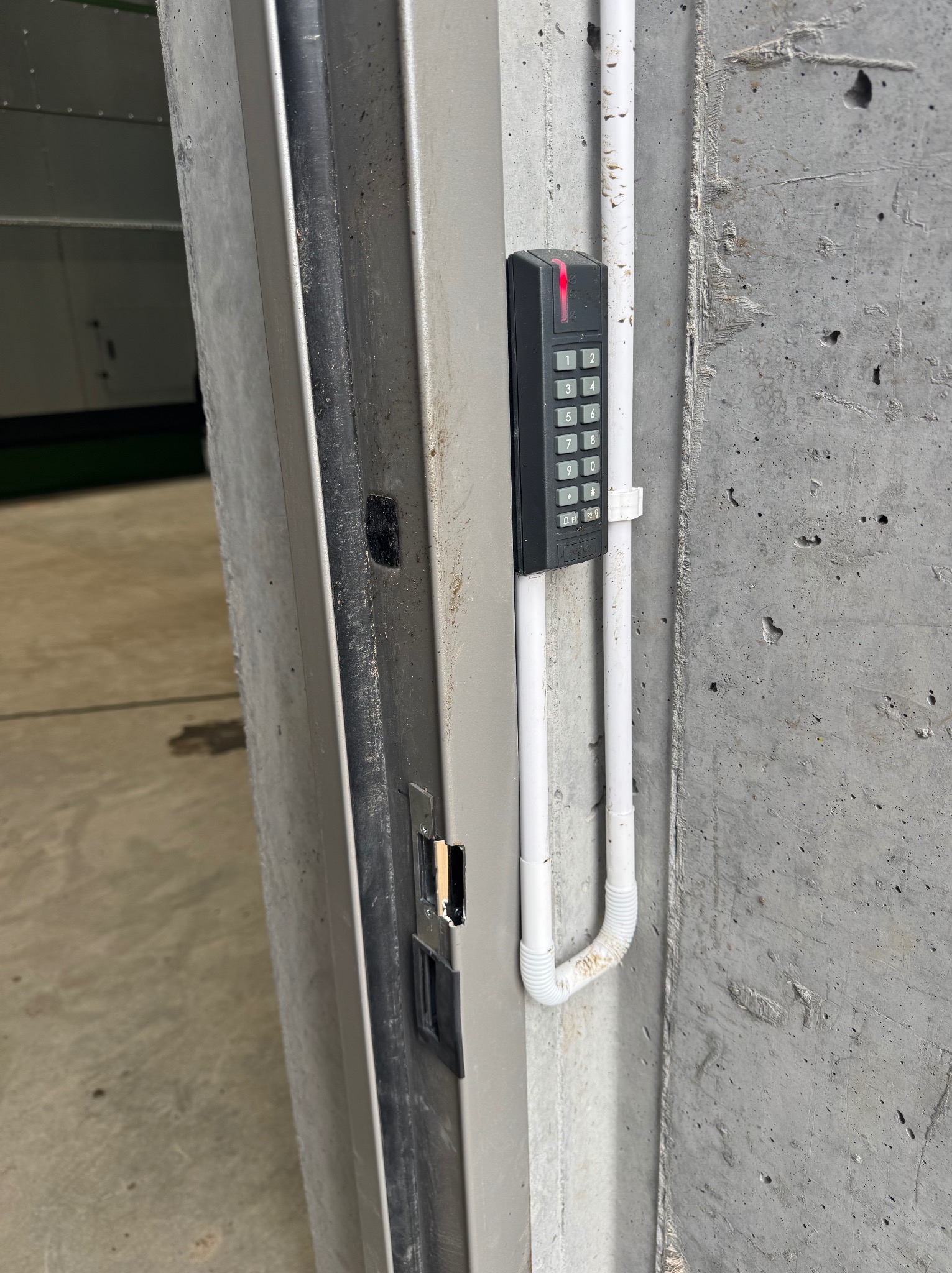 Zewnętrzny panel numeryczny domofonu z czerwoną diodą, zamontowany na surowej, betonowej ścianie obok metalowych drzwi z widocznym zamkiem.