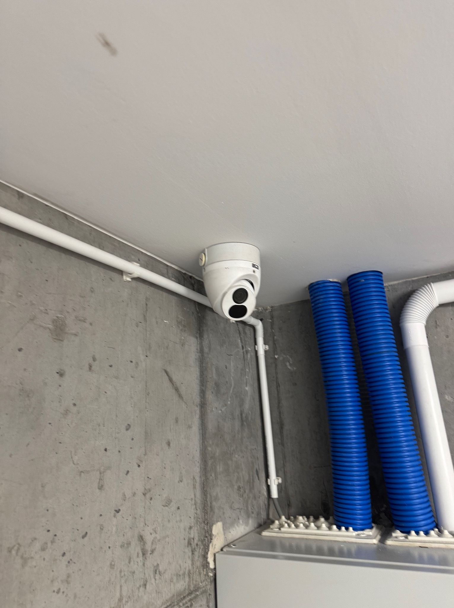 Kamera kopułkowa monitoringu zamontowana pod sufitem w surowym wnętrzu, widoczne rury instalacyjne w kolorze białym i niebieskim.