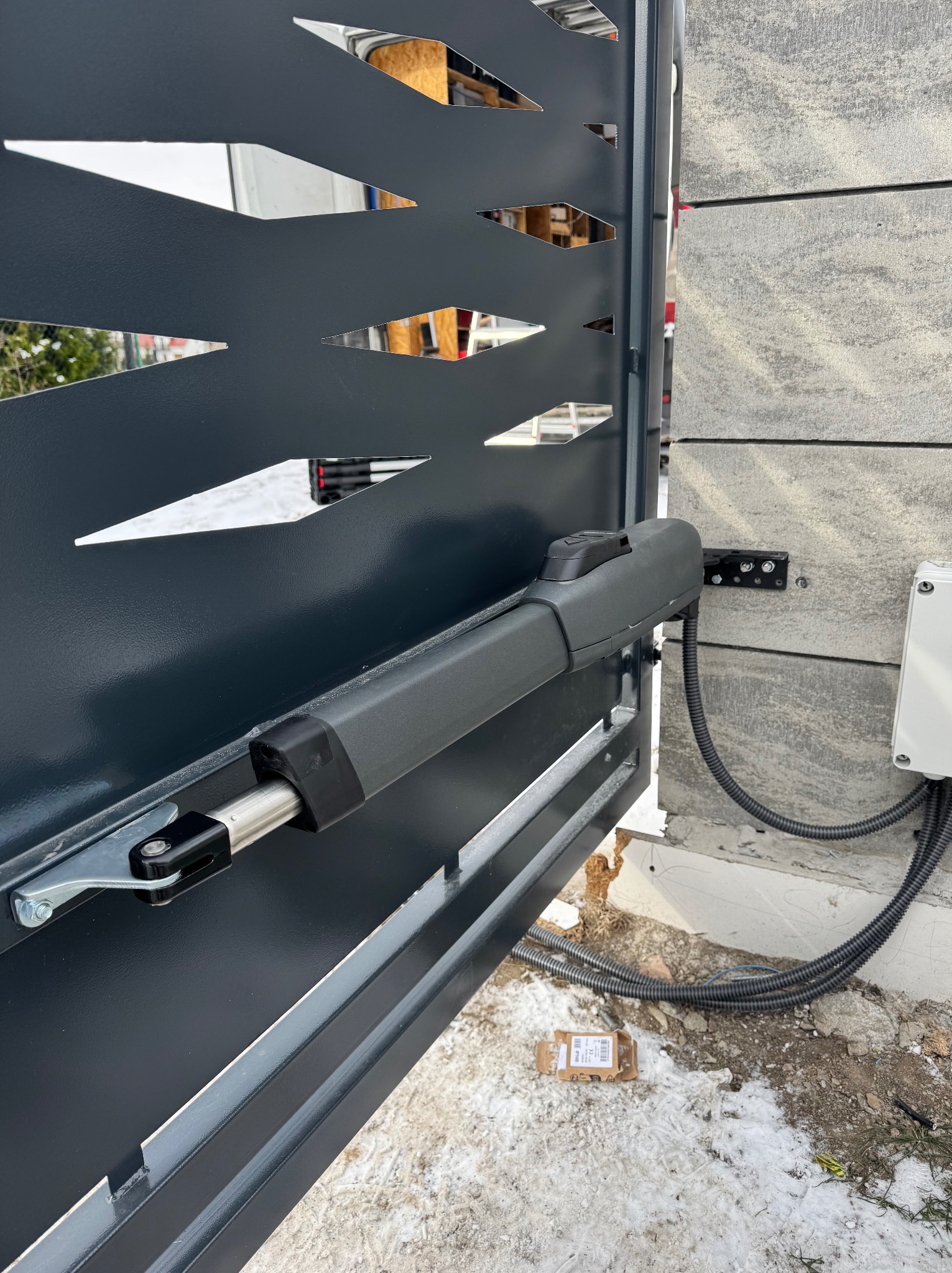 Szara, metalowa brama z automatyką, widoczny siłownik zamontowany na słupku z szarej, betonowej imitacji kamienia. Częściowo ośnieżone podłoże.