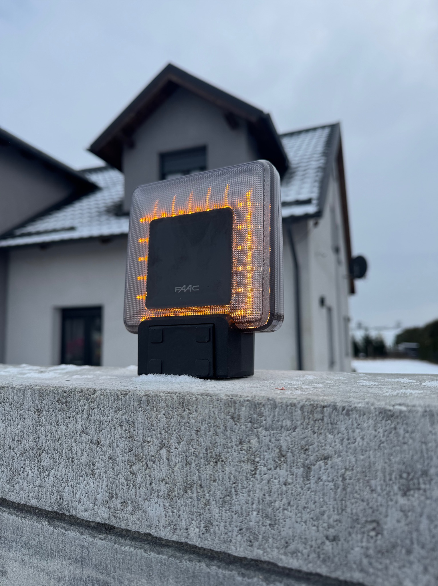 Lampa ostrzegawcza FAAC z pomarańczowym światłem, zamontowana na betonowym murku w zimowej scenerii, z domem w tle.