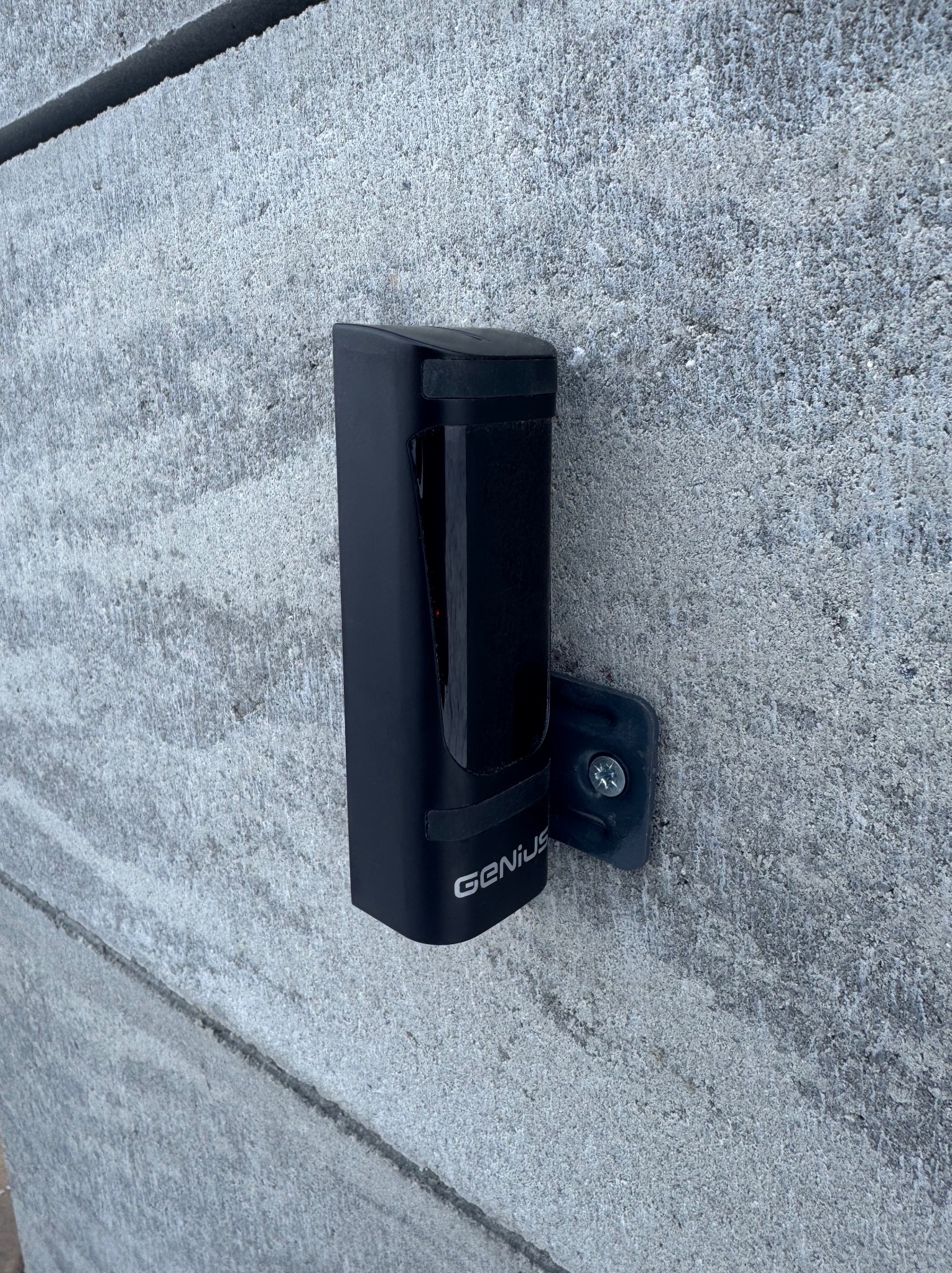 Czarny czujnik zbliżeniowy marki Genius zamontowany na szarej, betonowej ścianie z widoczną śrubą mocującą.