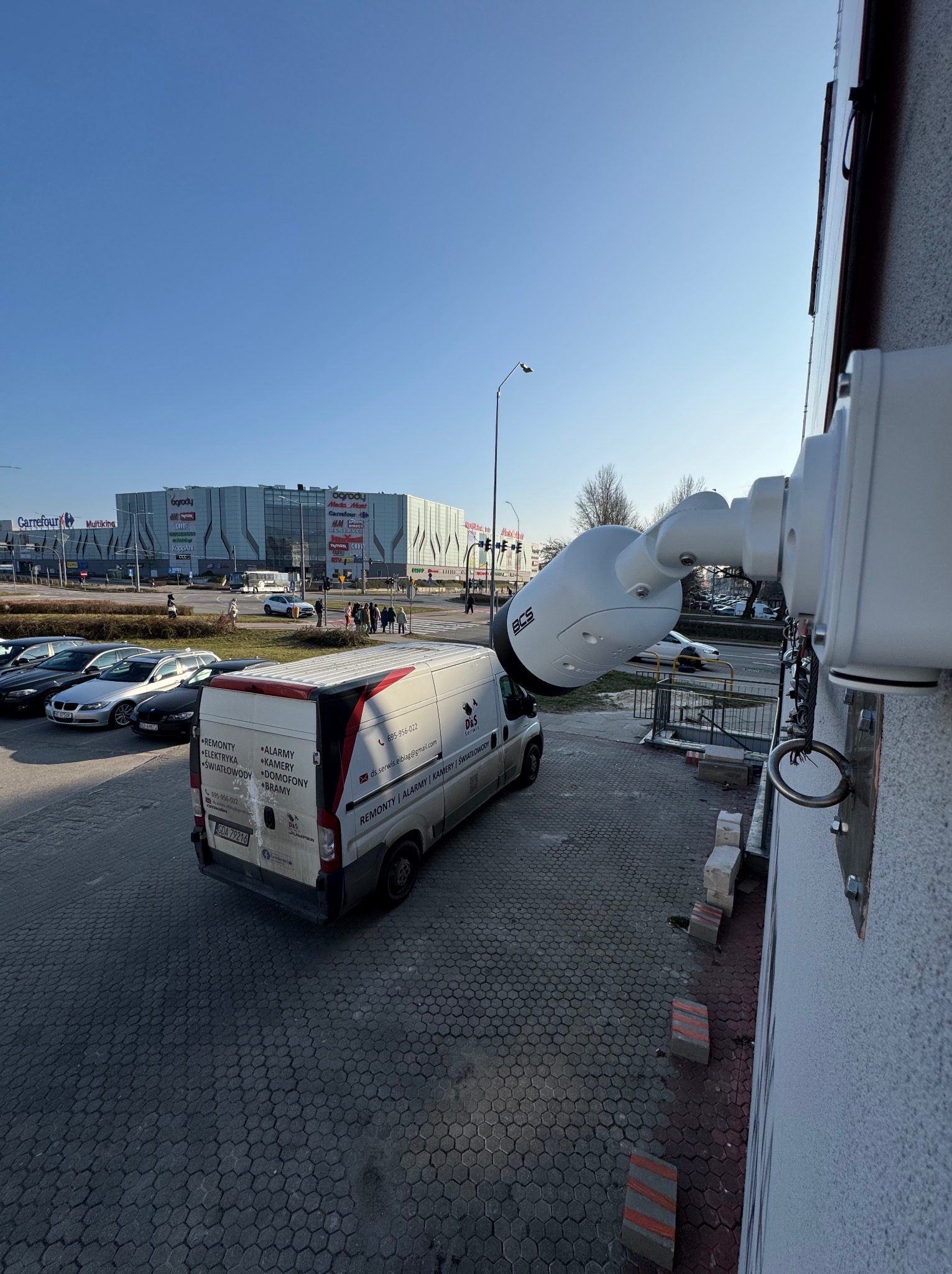 Kamera monitoringu BCS zamontowana na ścianie budynku, z widocznym w tle zaparkowanym busem firmy serwisowej i centrum handlowym.