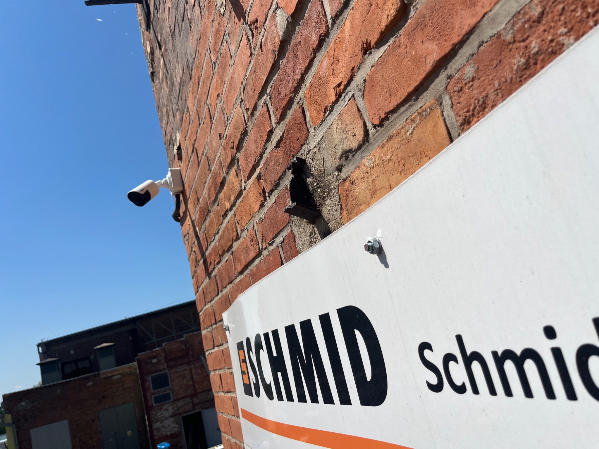 Kamera monitoringu zamontowana na ceglanej ścianie budynku, widoczna z dołu pod kątem, obok szyld z nazwą firmy.