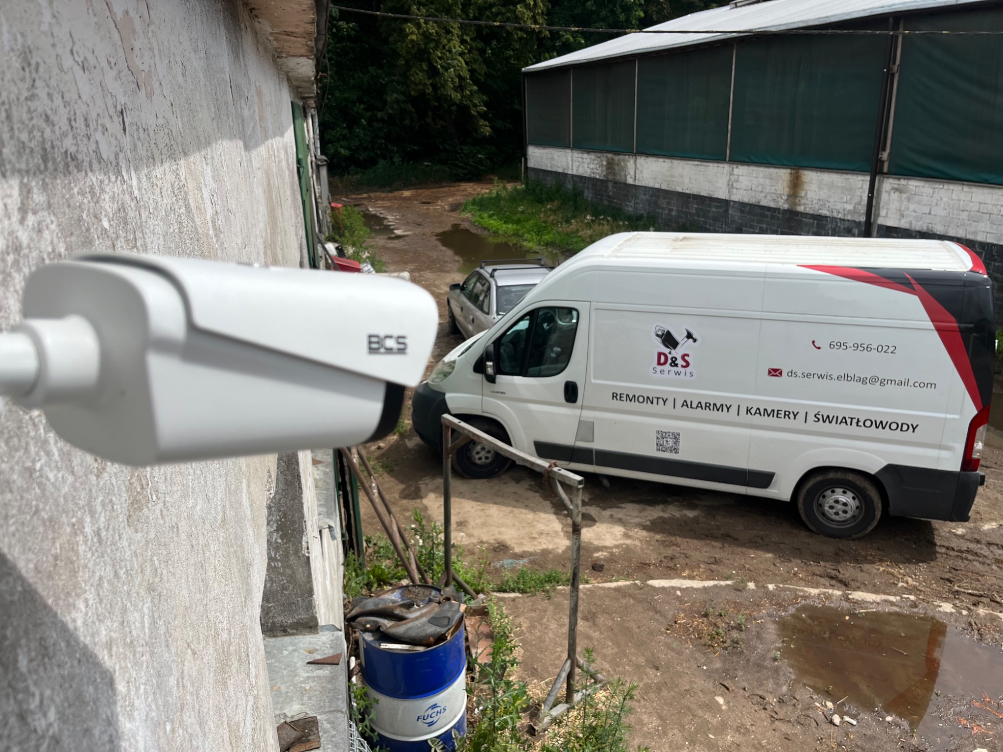 Zewnętrzna kamera monitoringu BCS zamontowana na szarej ścianie budynku, z widocznym samochodem serwisowym firmy D&S Serwis w tle na błotnistym podwórku.
