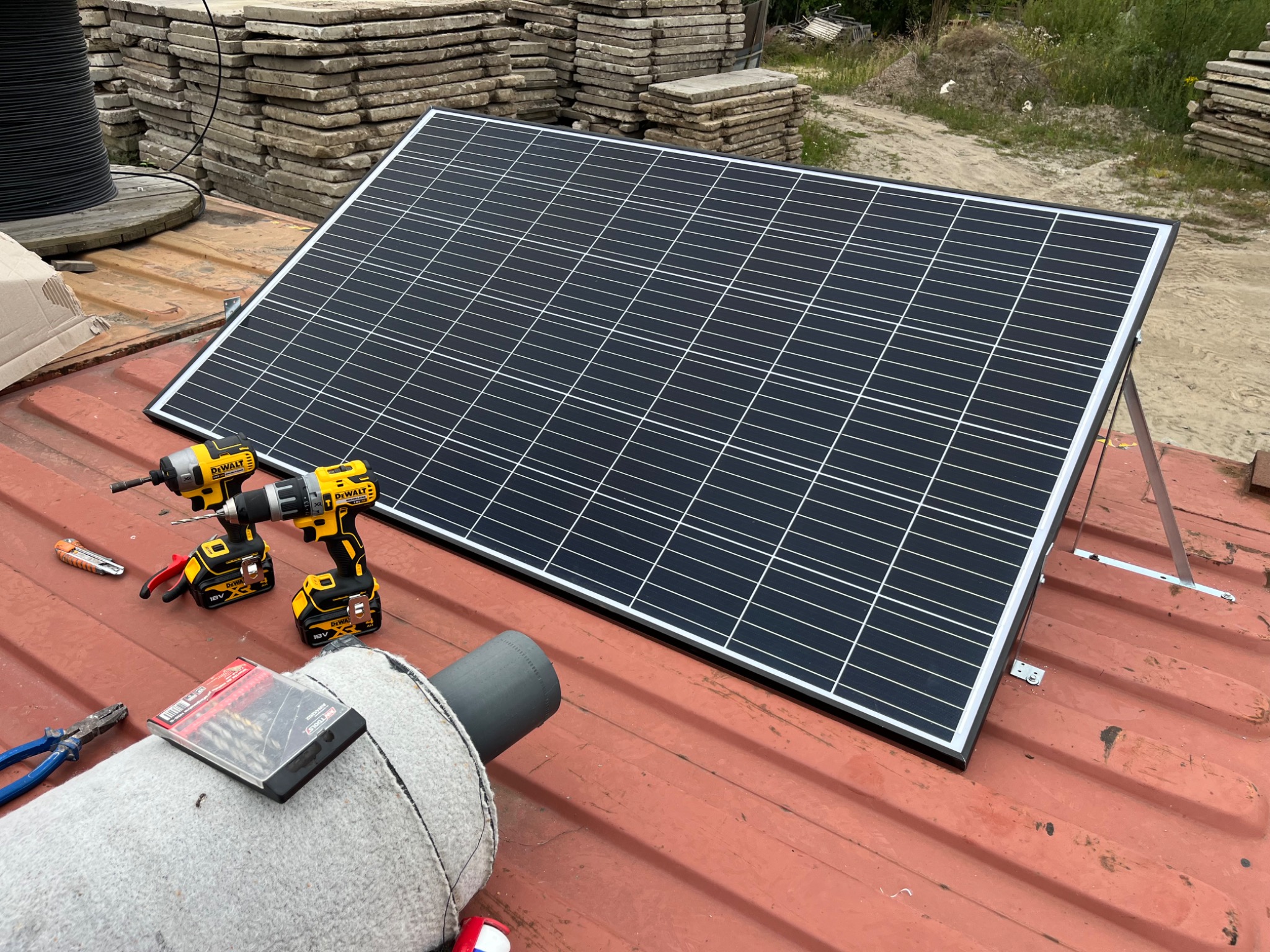 Panel fotowoltaiczny ustawiony na dachu z dwoma wkrętarkami DeWalt, narzędziami i materiałami przygotowanymi do montażu, z paletami w tle.