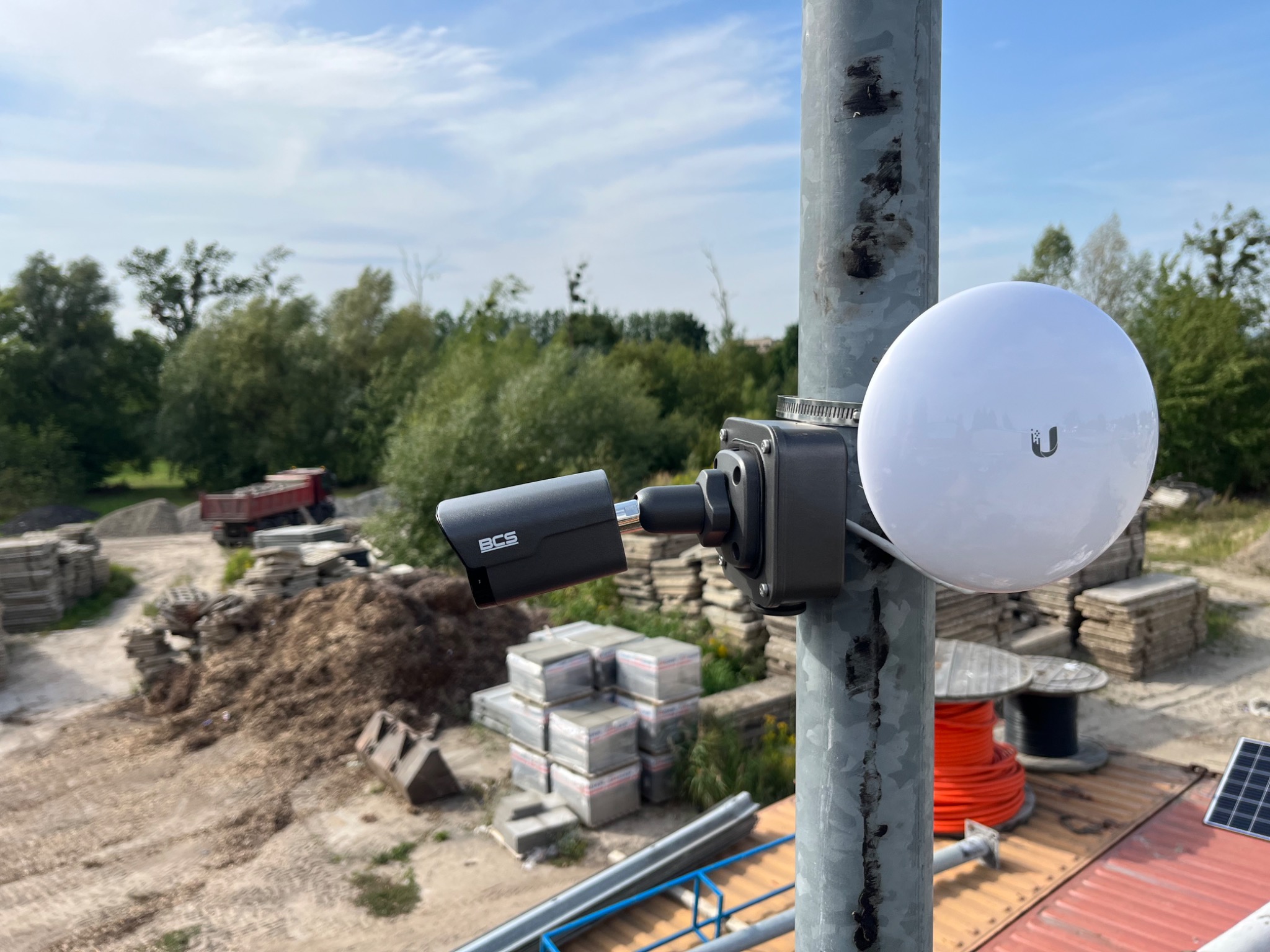 Kamera BCS i biała antena sferyczna zamontowane na szarej metalowej rurze na tle placu budowy z hałdą ziemi i stertą kostki brukowej.