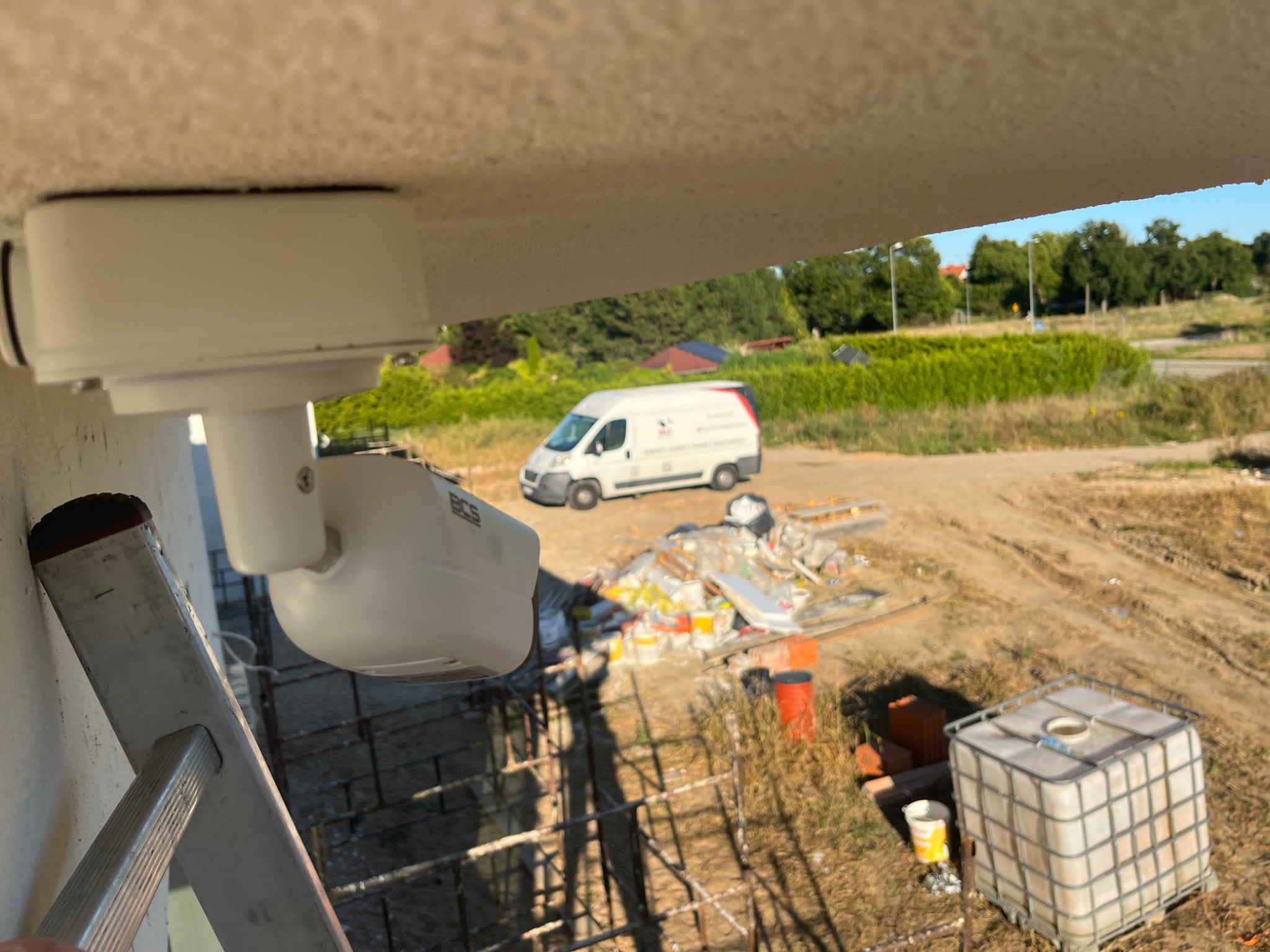 Kamera monitoringu BCS zamontowana pod okapem budynku, widoczna drabina i samochód dostawczy firmy na tle terenu budowy.