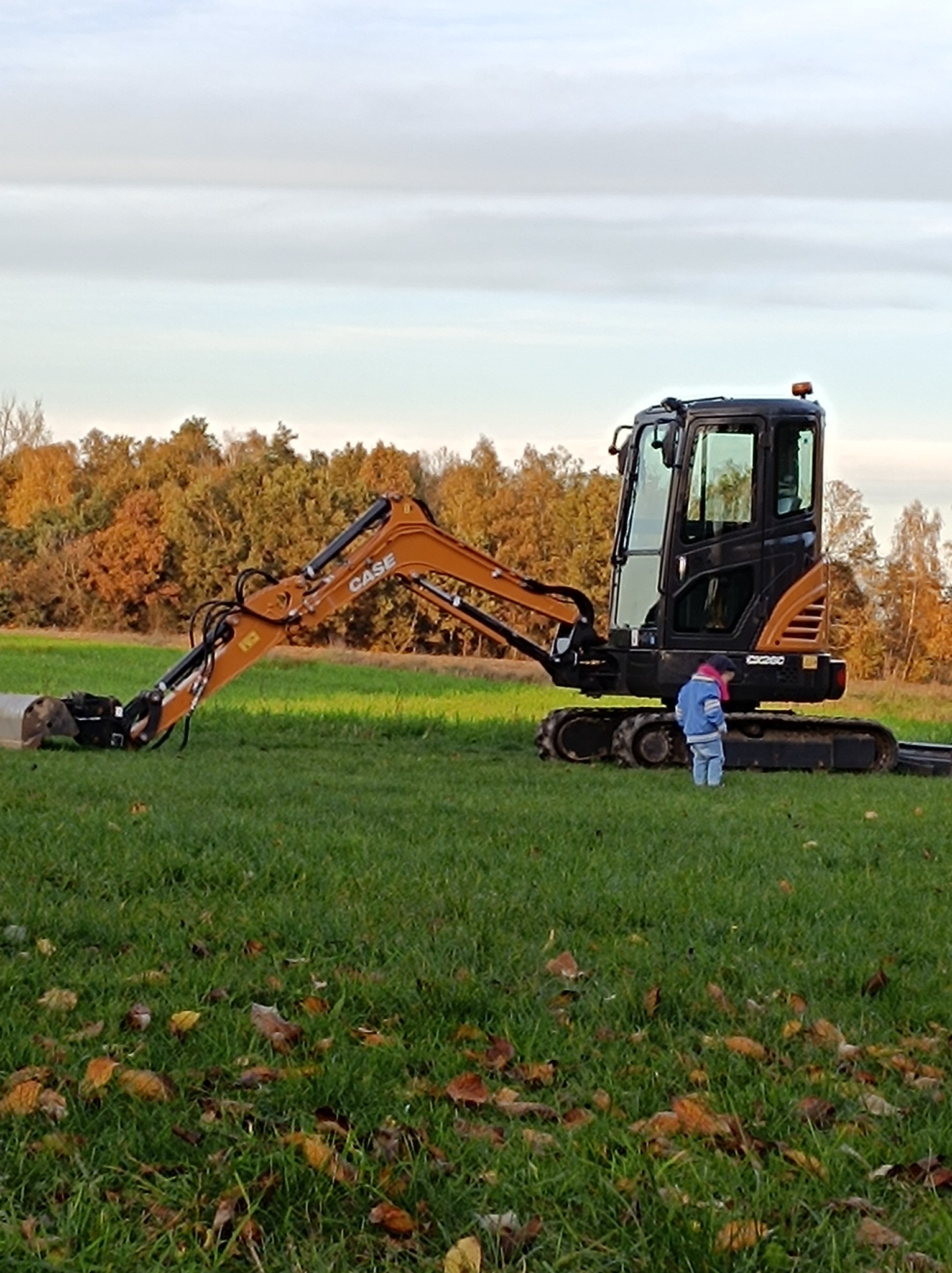 Mini koparka CASE na trawiastym terenie z jesiennymi drzewami w tle, obok stoi małe dziecko w niebieskiej kurtce.