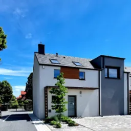 Nowoczesny budynek biurowy z białymi i szarymi ścianami, drewnianymi akcentami, szarym dachem i starannie zagospodarowanym terenem przed wejściem z młodym drzewkiem.