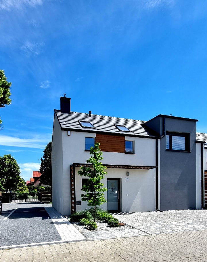 Nowoczesny budynek biurowy z białymi i szarymi ścianami, drewnianymi akcentami, szarym dachem i starannie zagospodarowanym terenem przed wejściem z młodym drzewkiem.