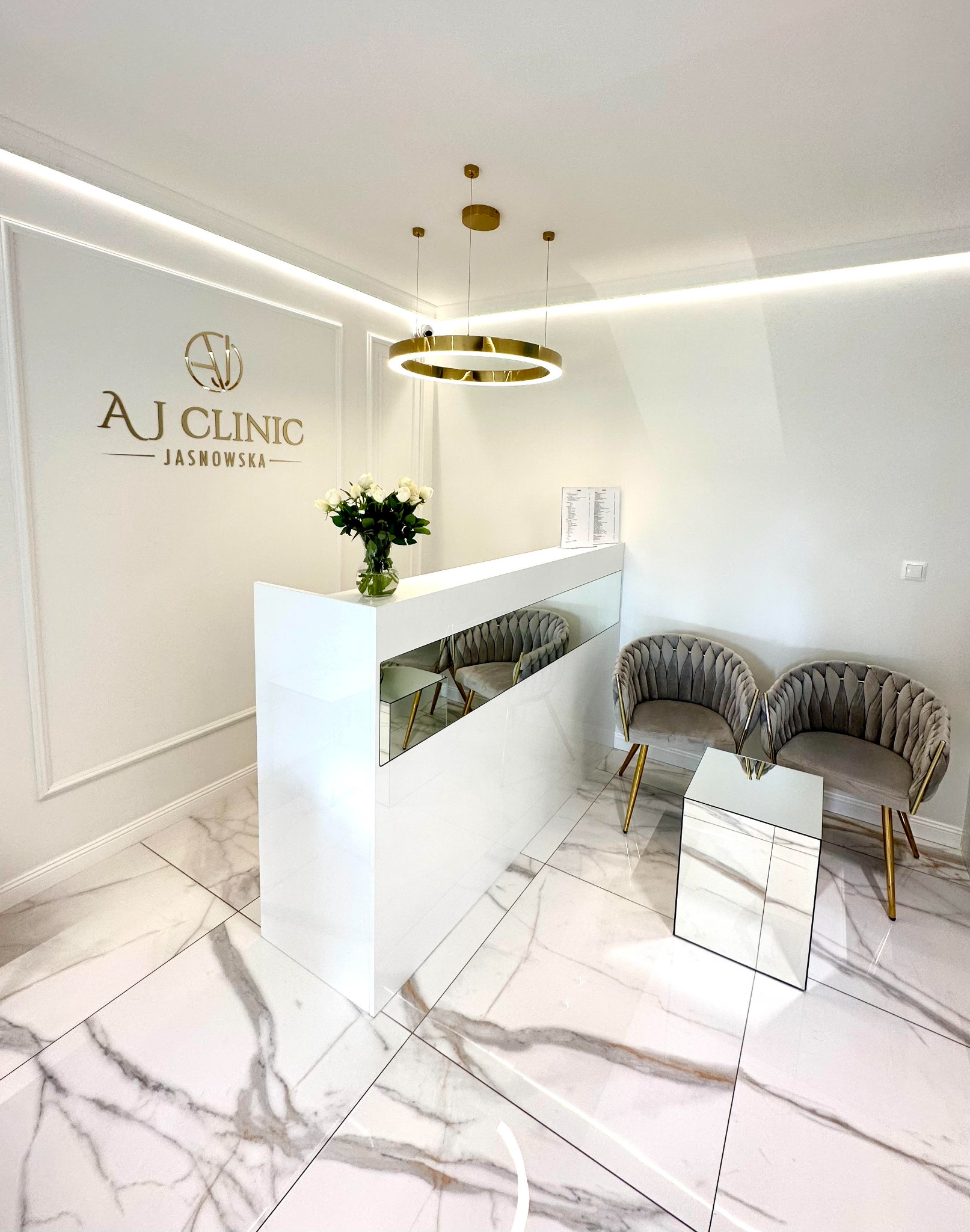 Nowoczesne wnętrze recepcji kliniki z białym biurkiem, kwiatami w wazonie, złotym oświetleniem i marmurową podłogą.