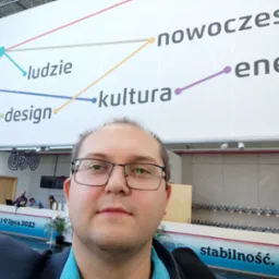 Mężczyzna w okularach robi selfie na tle ściany z napisem 'nowoczesność', 'ludzie', 'design', 'kultura', 'energia' i 'stabilność', w tle widoczne stanowisko obsługi.