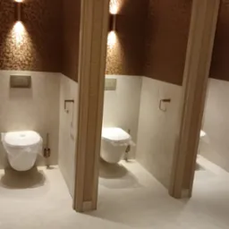 Rząd trzech kabin toaletowych z wiszącymi muszlami, zabezpieczonymi folią, w jasnej, nowoczesnej łazience wykończonej beżowymi płytkami i tapetą z geometrycznym wzorem, oświetlonych kinkietami.