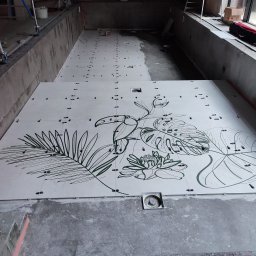 Firma ogólnobudowlana Marcin Jaśniewicz - Układanie jasnych płytek z motywem tropikalnym (liście, tukan) w nieckę basenową, widoczne poziomica i zwijak kabla, w tle drabina i okno.