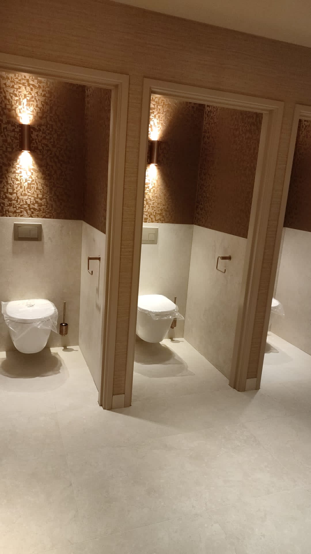 Rząd trzech kabin toaletowych z wiszącymi muszlami, zabezpieczonymi folią, w jasnej, nowoczesnej łazience wykończonej beżowymi płytkami i tapetą z geometrycznym wzorem, oświetlonych kinkietami.