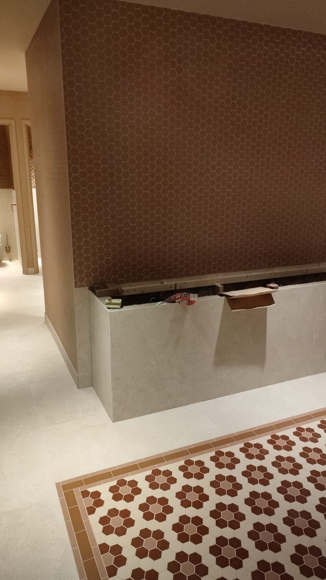 Ściana wykończona tapetą w brązowe heksagony, z widocznym fragmentem zabudowy z płyt, częściowo otwartej, ukazującej wnętrze z elementami instalacji i materiałów budowlanych; na podłodze wzorzysta,...