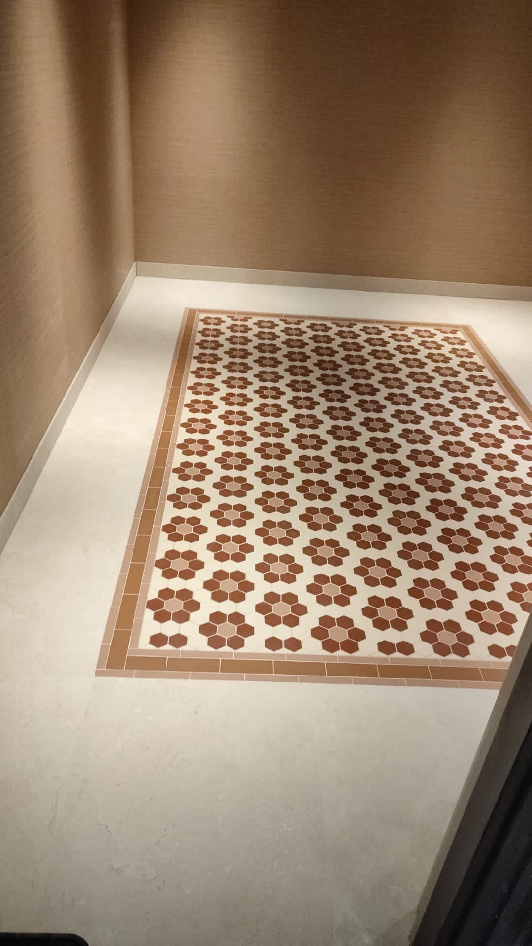 Wzorzysta, brązowo-biała podłoga z płytek w kształcie kwiatów, otoczona beżowymi ścianami i jasnymi płytkami podłogowymi.