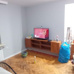 Usługi Remontowo-Wykończeniowe JAK-BUD Kacper Jakubiak - Pokój w trakcie sprzątania po remoncie: odkurzacz, wiadro, śmieci w worku, zabezpieczona podłoga parkietowa, szare ściany, meble pod telewizorem.