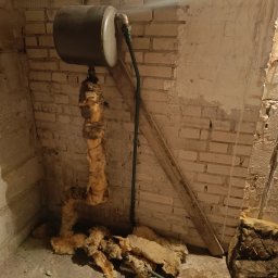 Usługi Remontowo-Wykończeniowe JAK-BUD Kacper Jakubiak - Zdjęcie przedstawia stary, zaizolowany zbiornik na wodę w piwnicy z odsłoniętą cegłą i fragmentami izolacji na podłodze, ukazując stan zaniedbania i potrzebę remontu.