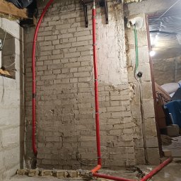 Usługi Remontowo-Wykończeniowe JAK-BUD Kacper Jakubiak - Montaż instalacji centralnego ogrzewania w piwnicy z odsłoniętą cegłą, widoczne czerwone rury, zbiornik wyrównawczy i taśma miernicza na podłodze.