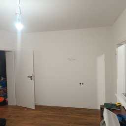 Usługi Remontowo-Wykończeniowe JAK-BUD Kacper Jakubiak - Pokój przed remontem: białe ściany z wystającymi kablami, surowa żarówka, drewniana podłoga i widoczne narzędzia oraz materiały budowlane na meblu w rogu.