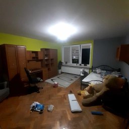 Usługi Remontowo-Wykończeniowe JAK-BUD Kacper Jakubiak - Pokój z drewnianą podłogą, meblami i ścianami w dwóch kolorach: zielonym i szarym. Na podłodze leżą przedmioty. W tle widać okno i grzejnik.