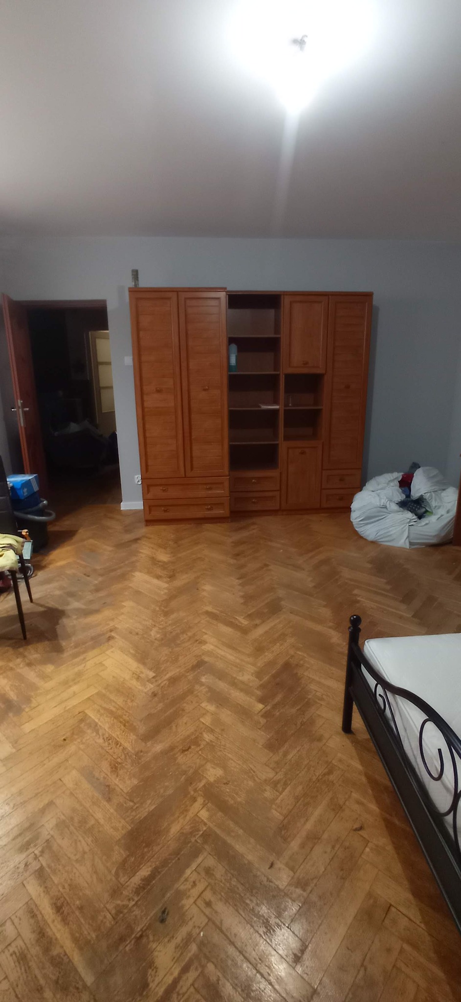 Pokój z parkietem ułożonym w jodełkę, drewnianą szafą z półkami i szufladami oraz częściowo widocznym łóżkiem z metalową ramą, w tle otwarte drzwi do kolejnego pomieszczenia.