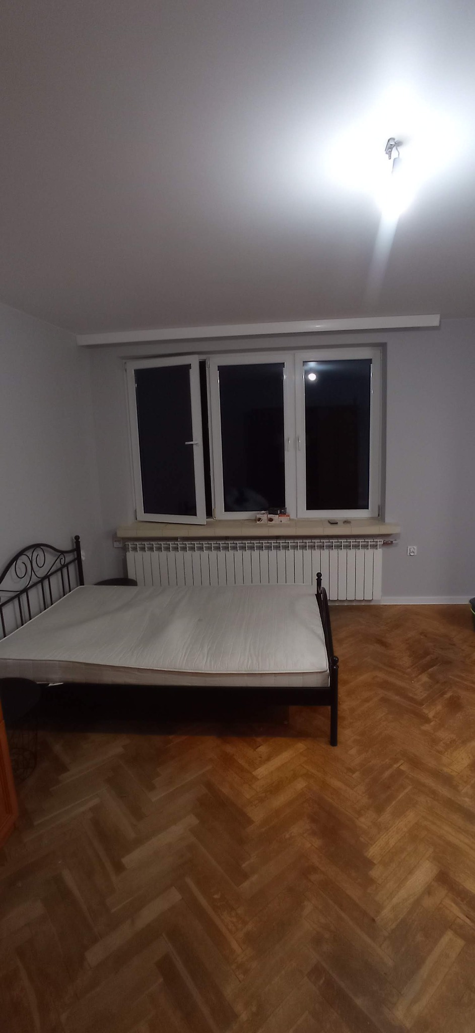 Pokój z parkietem ułożonym w jodełkę, metalowe łóżko z materacem bez pościeli, okno z grzejnikiem pod spodem, ściany pomalowane na szaro.
