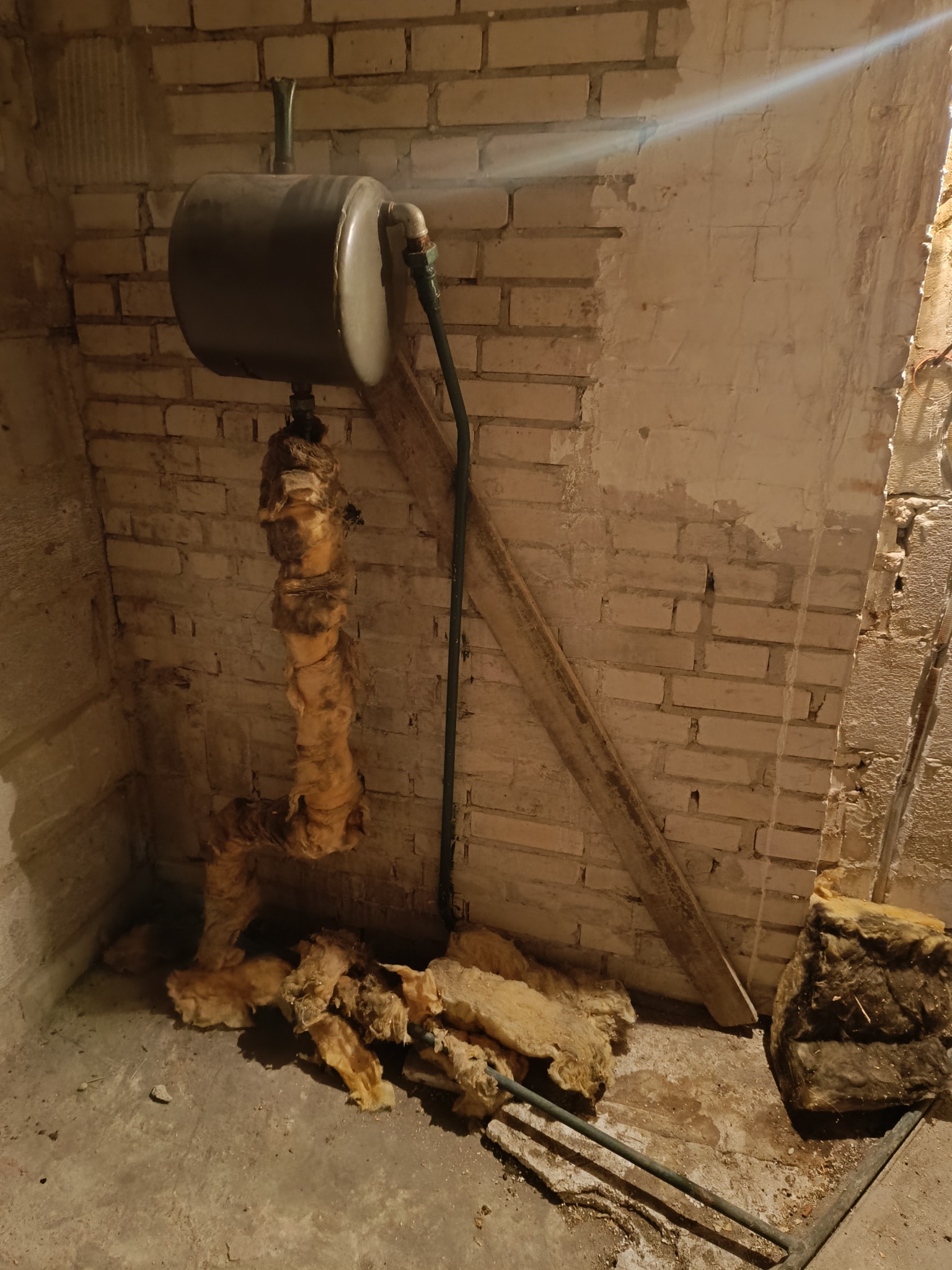 Niedokończone ocieplenie rur w piwnicy z odsłoniętym murem z cegły i fragmentami izolacji na podłodze.