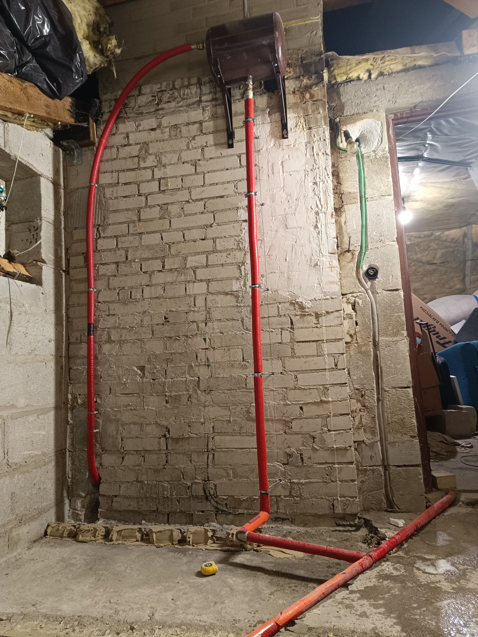 Montaż instalacji centralnego ogrzewania z rur w kolorze czerwonym na tle ściany z cegły w trakcie remontu, widoczny zbiornik wyrównawczy i żółta miara zwijana.