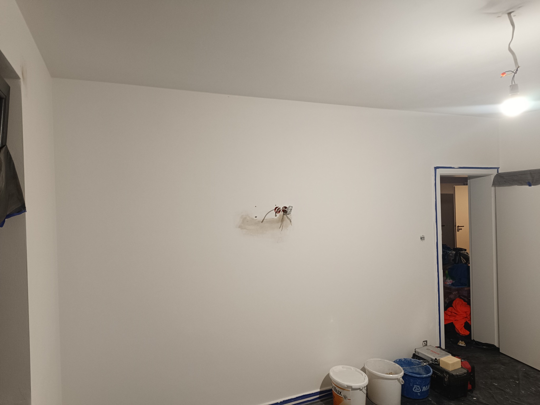 Wnętrze pokoju w trakcie odświeżania: świeżo pomalowana biała ściana z widocznym otworem na gniazdko elektryczne z wystającymi kablami, zabezpieczona taśmą malarską podłoga i futryna drzwi,...